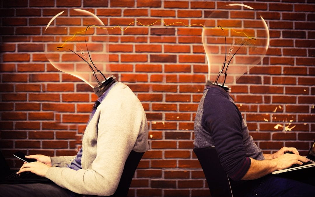 Image of two people with light bulbs as heads sitting back to back at computers. Their heads are connected by a line of electricity, symbolizing inspiration.