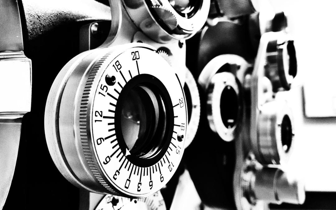 Black and white close-up photograph of an optometrist’s phoropter, an instrument used for eye examinations. The image prominently features several large, circular dials with numbers and lines, which are used to change lenses to test a patient’s vision. The numbers on the dials range from 0 to around 20, including the different lens strengths or prescriptions. The metal parts of the phoropter have a reflective surface, creating a contrast between light and shadow in the image.